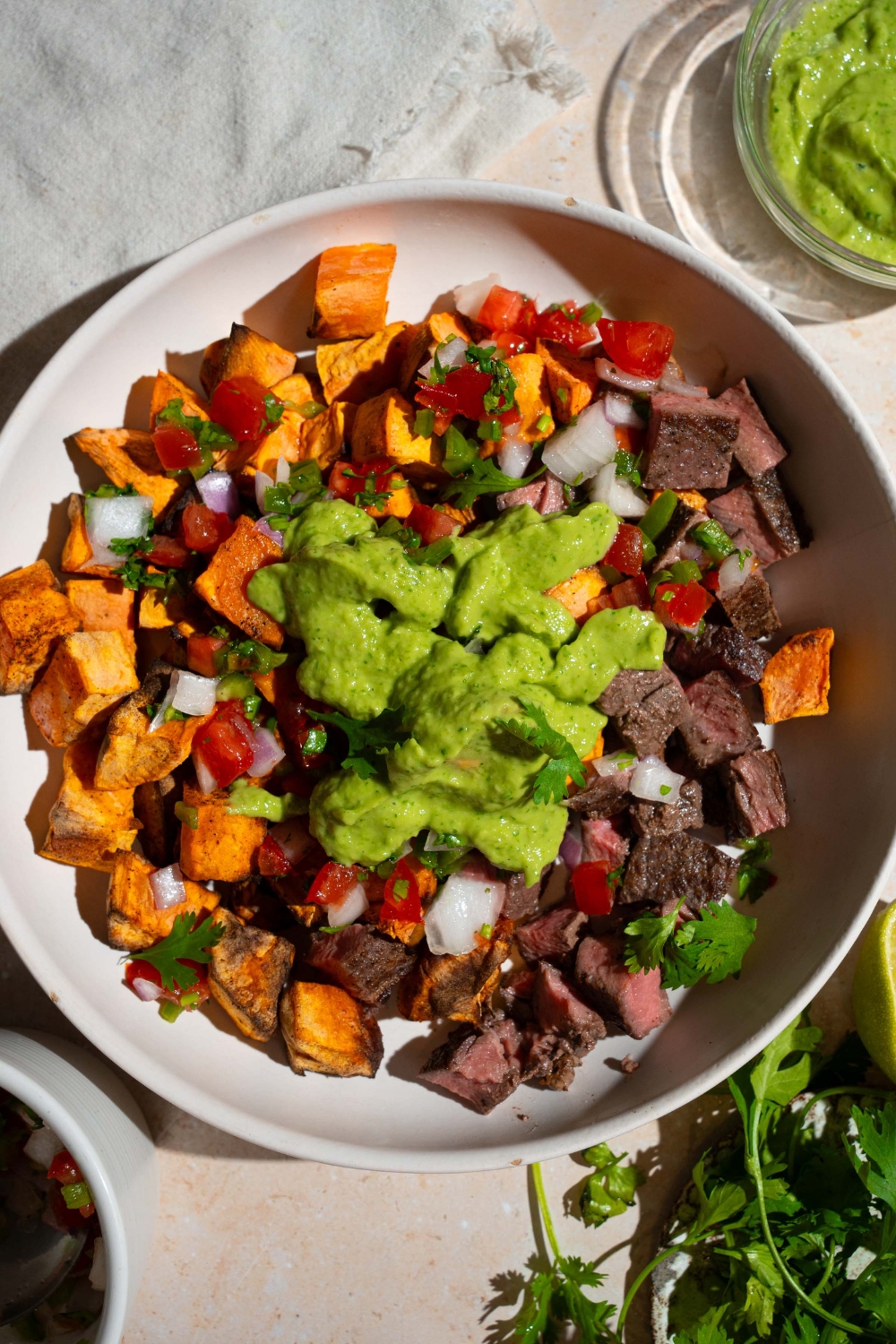 A sweet potato steak bowl topped with pico de gallo and avocado cilantro lime dressing. The bowl is on a tan counter with fresh cilantro with a small bowl of dressing.
