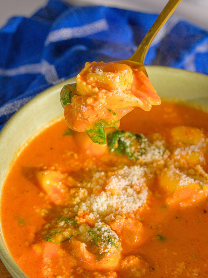 A spoon with a tortellini on it being held above a bowl of tortellini tomato soup.