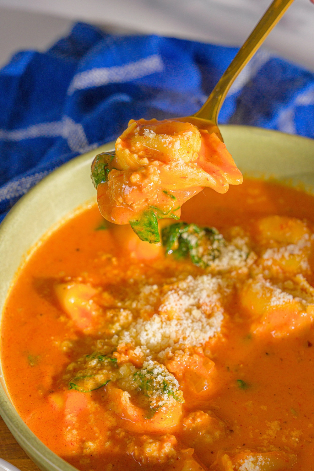 A spoon with a tortellini on it being held above a bowl of tortellini tomato soup.