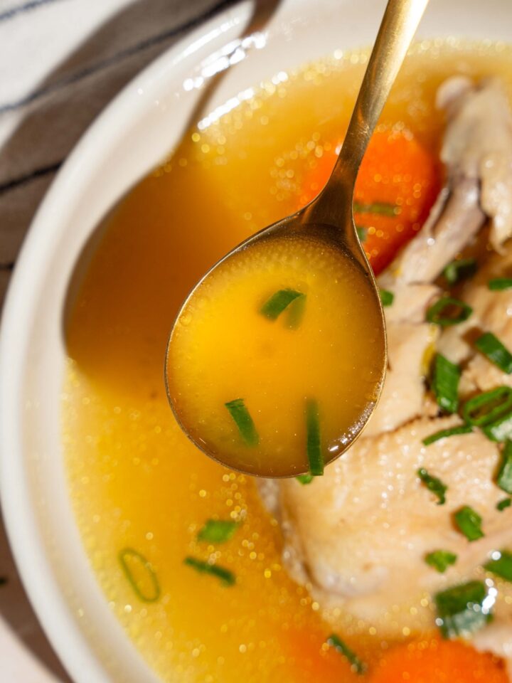 A spoon of waterless chicken soup broth being held above a bowl that is filled with the no water chicken soup.
