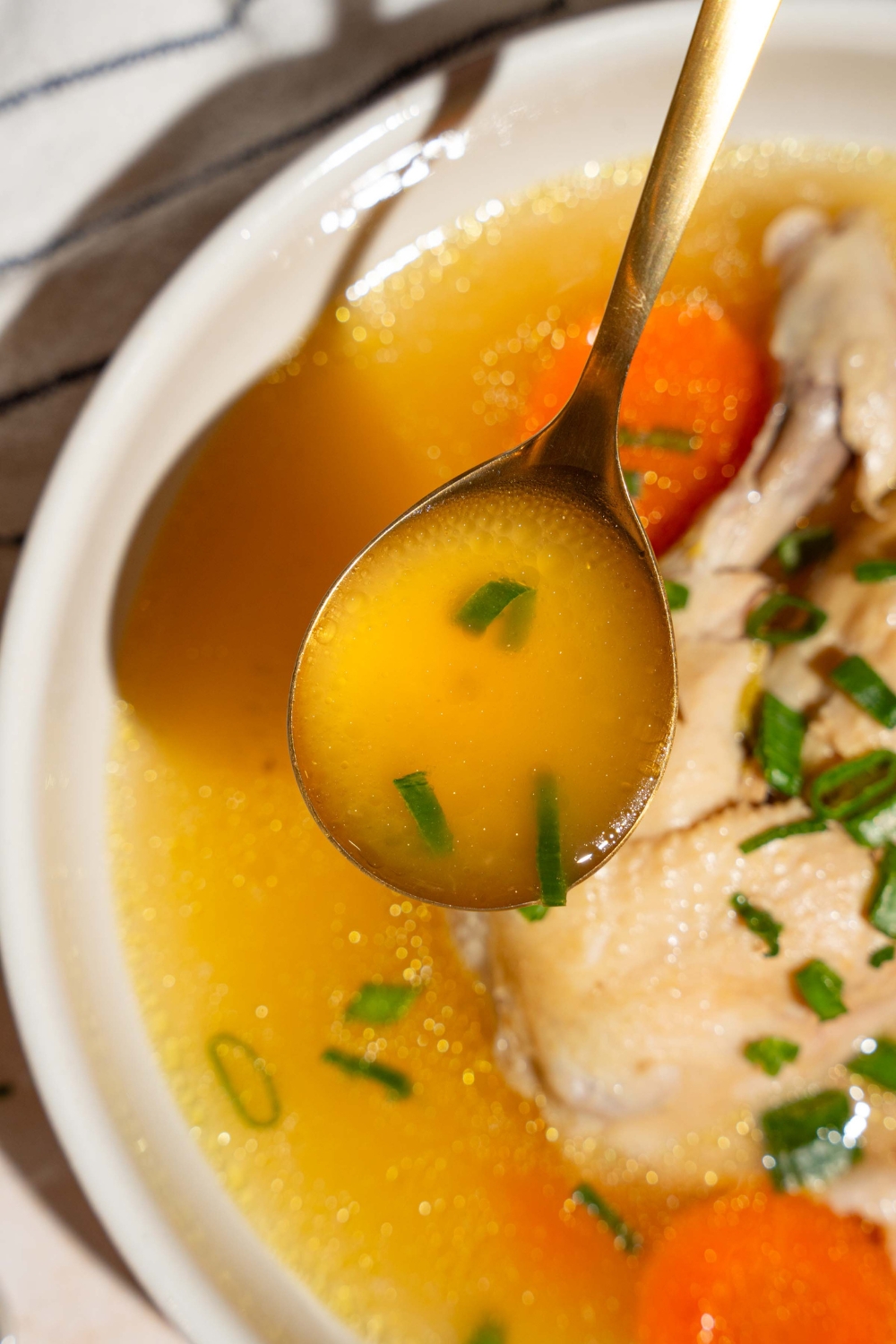 A spoon of waterless chicken soup broth being held above a bowl that is filled with the no water chicken soup.